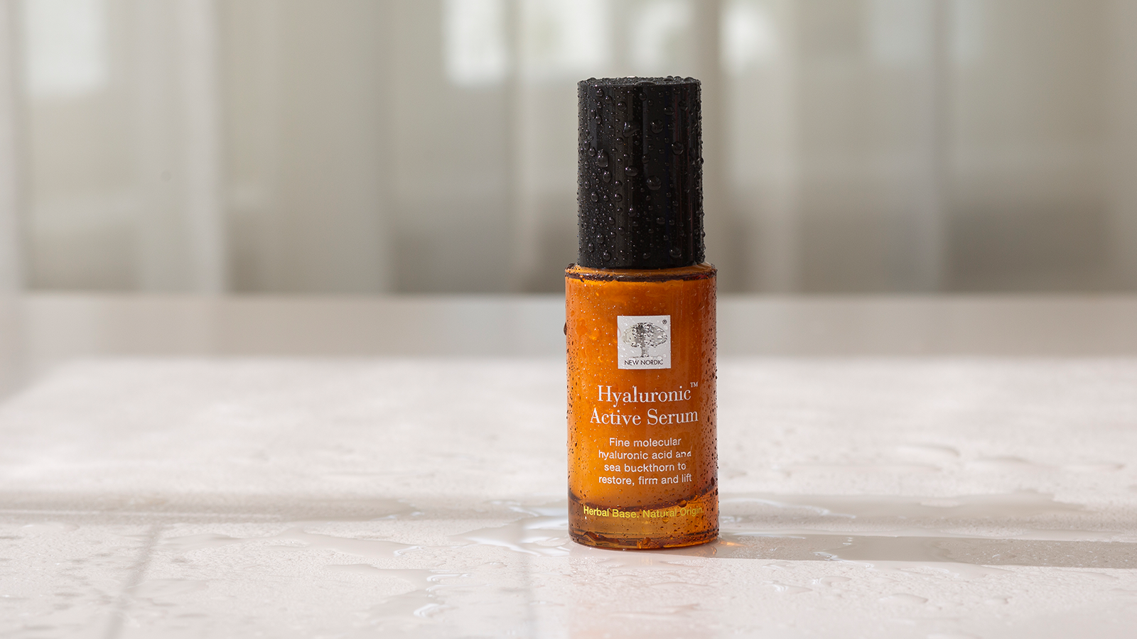 Fugtboosteren over dem alle 14 New Nordic Hyaluronic Active Serum™ i ravfarvet flaske med vanddråber på lys baggrund.