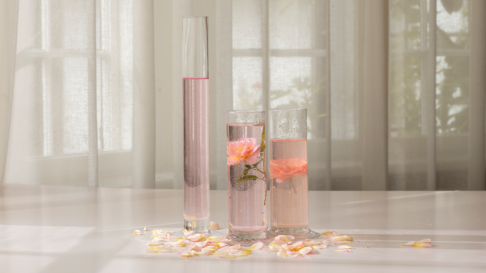 Tre glasvaser med lyserødt vand og blomster, omgivet af spredte lyserøde blomsterblade på et lyst bord.
