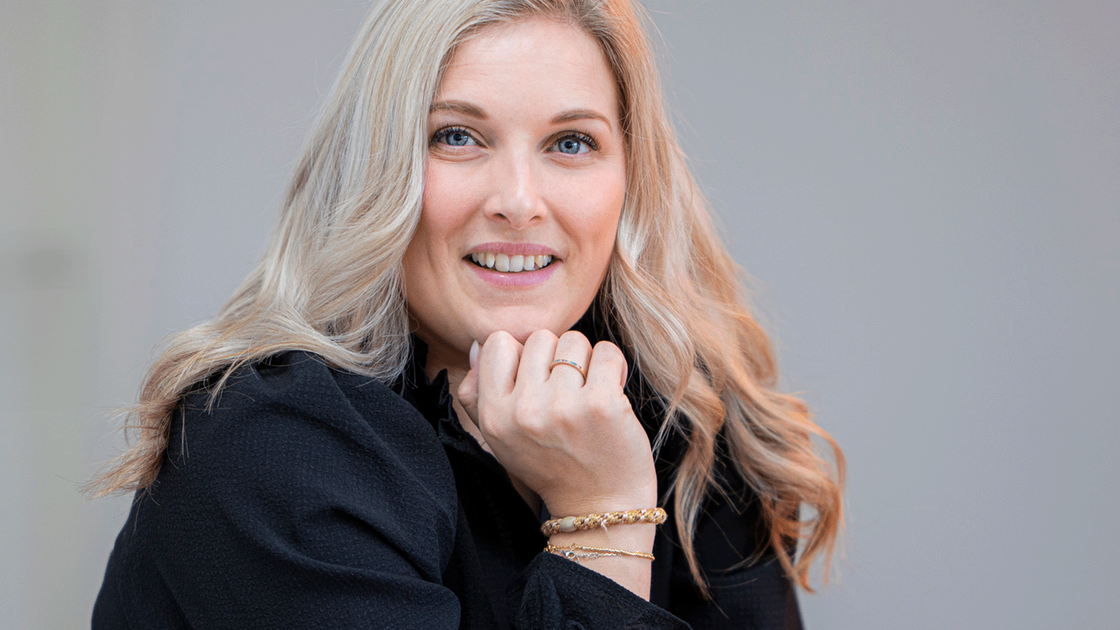 Smilende kvinde med lyst hår ser direkte mod kameraet med hånden under hagen.