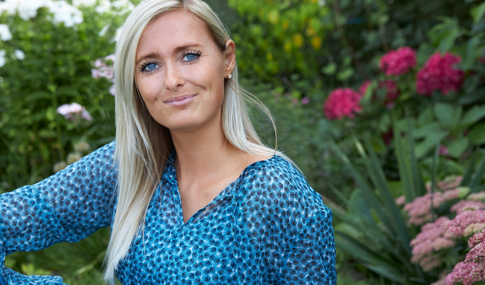 Smilende kvinde med lyst hår sidder udendørs i en blomstrende have.
